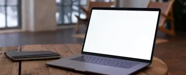 Portátil abierto con la pantalla en blanco sobre una mesa redonda de madera, junto a una libreta