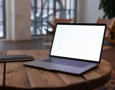 Portátil abierto con la pantalla en blanco sobre una mesa redonda de madera, junto a una libreta