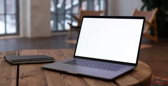 Portátil abierto con la pantalla en blanco sobre una mesa redonda de madera, junto a una libreta