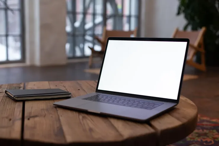 Portátil abierto con la pantalla en blanco sobre una mesa redonda de madera, junto a una libreta